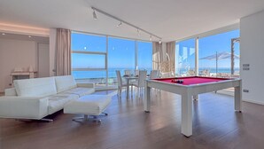 A modern living room with a pool table, white sofa, dining table, and chairs, large windows with a view of the sea.