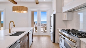 Una cocina moderna con armarios blancos, electrodomésticos de acero inoxidable y vista a la playa a través de una puerta abierta.