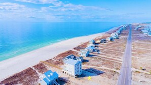 An aerial view of a coastal area with a long stretch of beach, a row of buildings, and a clear blue sky.