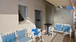 Un pequeño espacio al aire libre con dos sillas blancas y una mesa, una ventana con cortina y una puerta con persiana metálica.