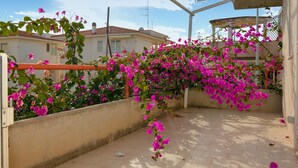 Un balcón con flores de buganvilla rosadas, piso de baldosas y vista a casas al fondo.