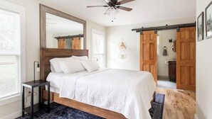 Un dormitorio con cama, ventilador de techo, espejo grande, puertas de madera y una pequeña mesa.