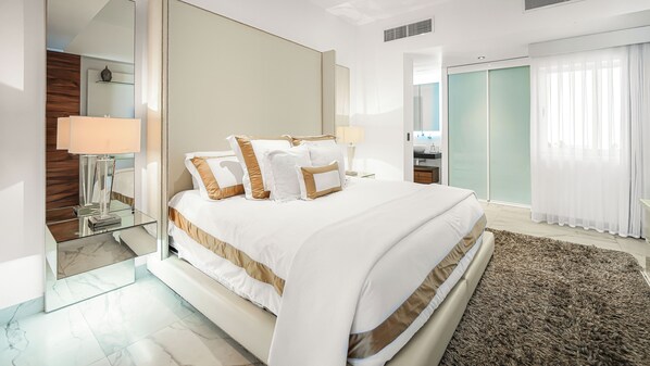 A modern hotel bedroom with a large bed, a glass bedside table, a mirror, and a view of the bathroom.