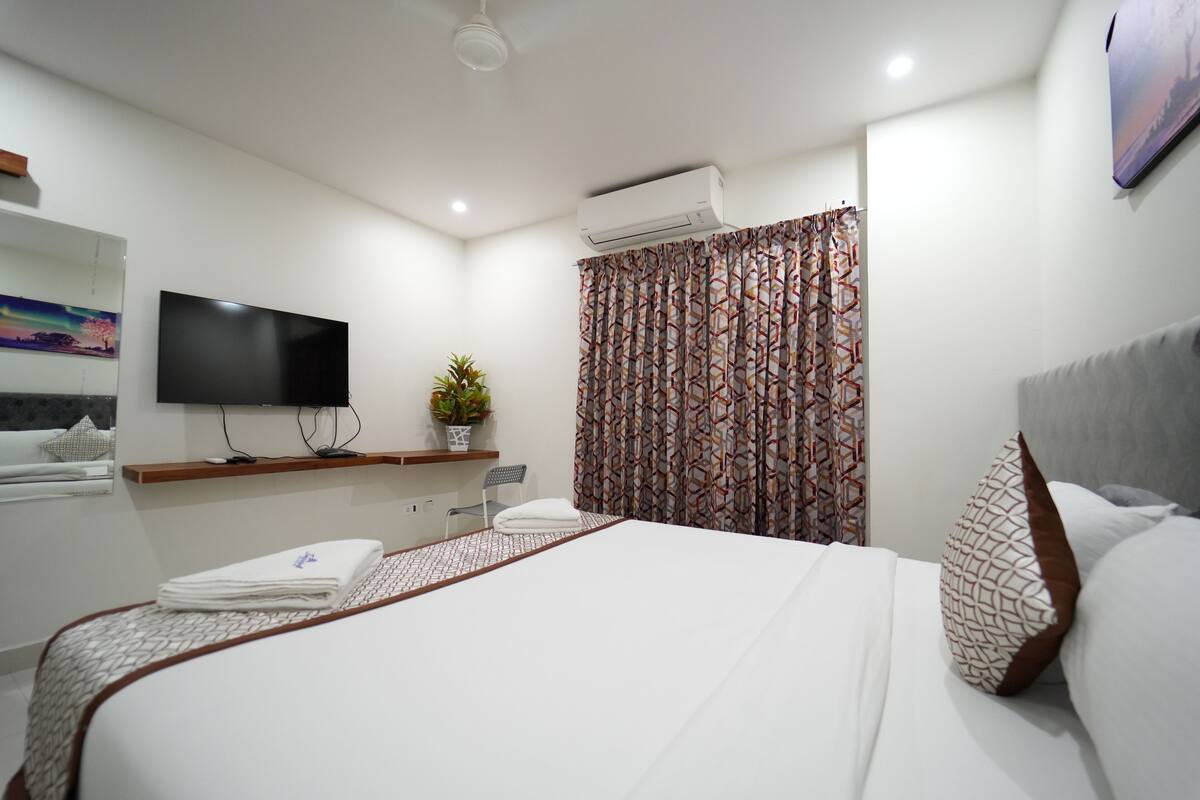 Ein modernes Hotelzimmer mit einem großen Bett, einem flachbildfernseher an der Wand, einer Klimaanlage, einem gemusterten Vorhang und einer kleinen Topfpflanze.