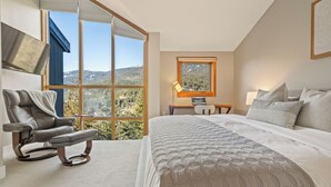 A bedroom with a large bed, a reclining chair, a desk, and a view of a mountainous landscape through large windows.