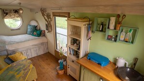 Una habitación acogedora con una cama, una mesa de comedor de madera, una pequeña cocina con un fregadero y estantes, y una ventana con vistas a la vegetación.