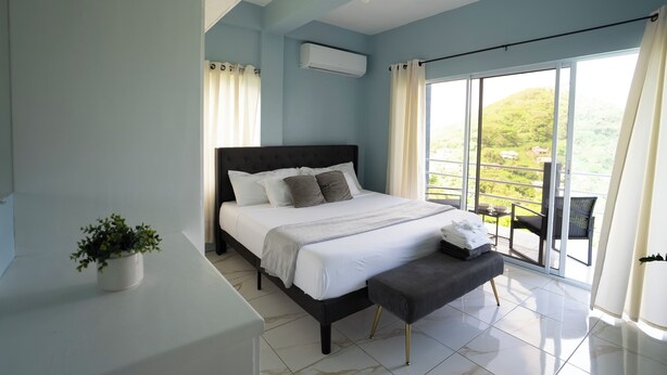 A bedroom with a bed, a small table, a bench, a potted plant, and a large window with a view of greenery.