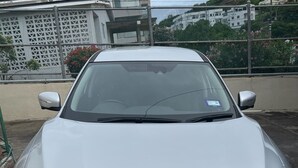 A silver Nissan car parked in a lot with a fence and buildings in the background.