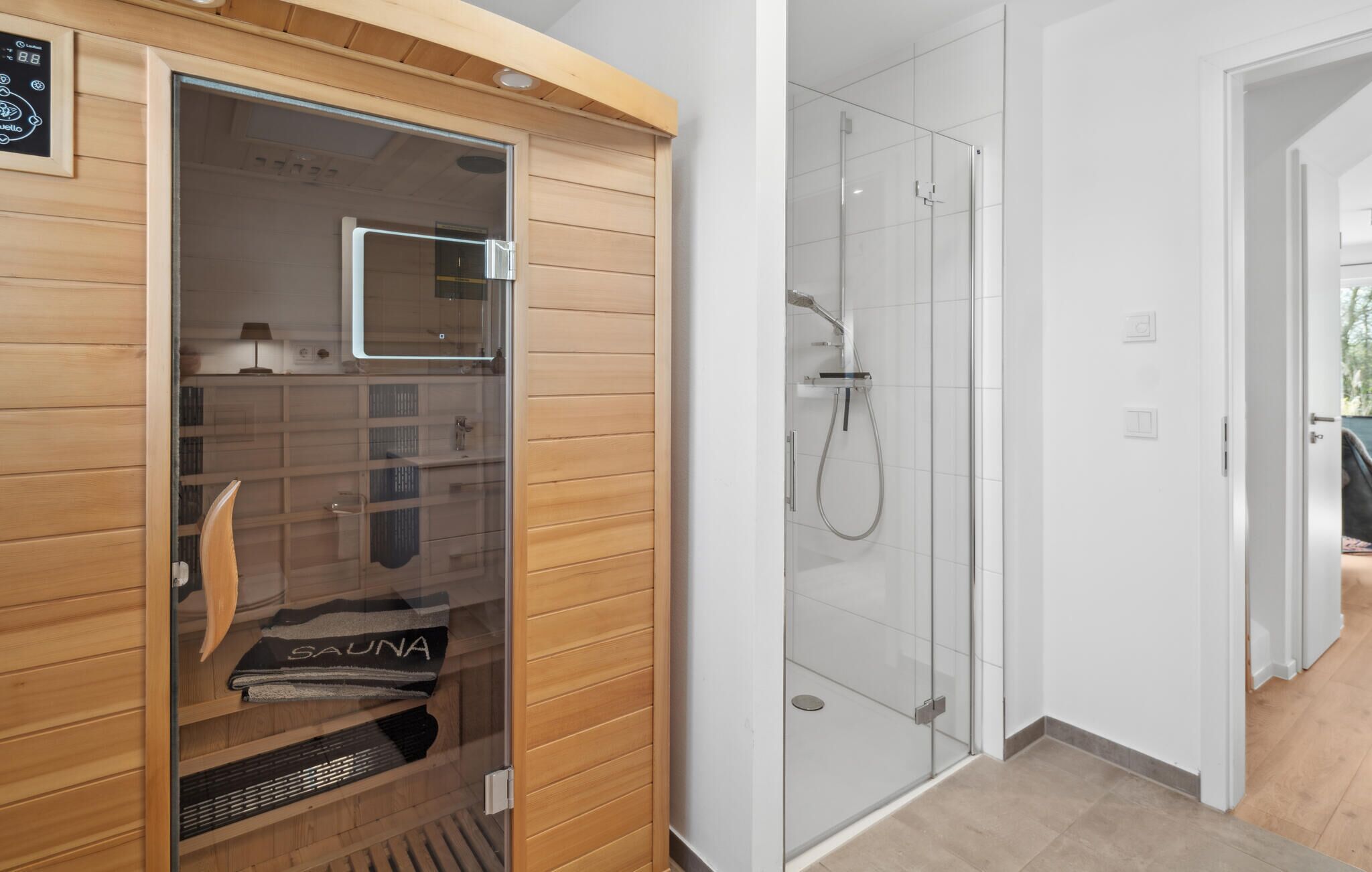 Eine Sauna mit Holzeinrichtung und Glastüren, ein Duschbereich mit Glaswänden sowie ein sichtbares Bedienfeld.