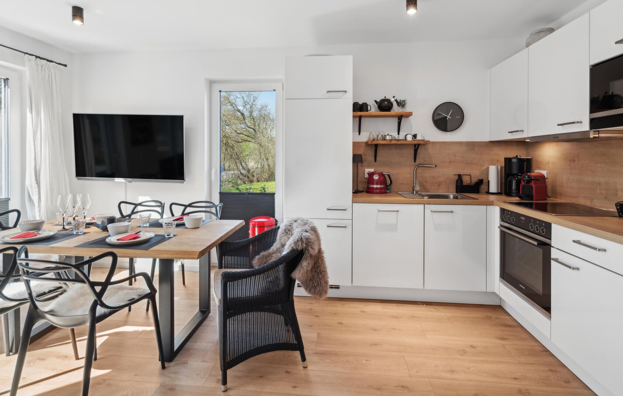 A modern kitchen with white cabinets, a wooden countertop, and a built-in oven. There is a dining area with a table and chairs, and a TV mounted on the wall.