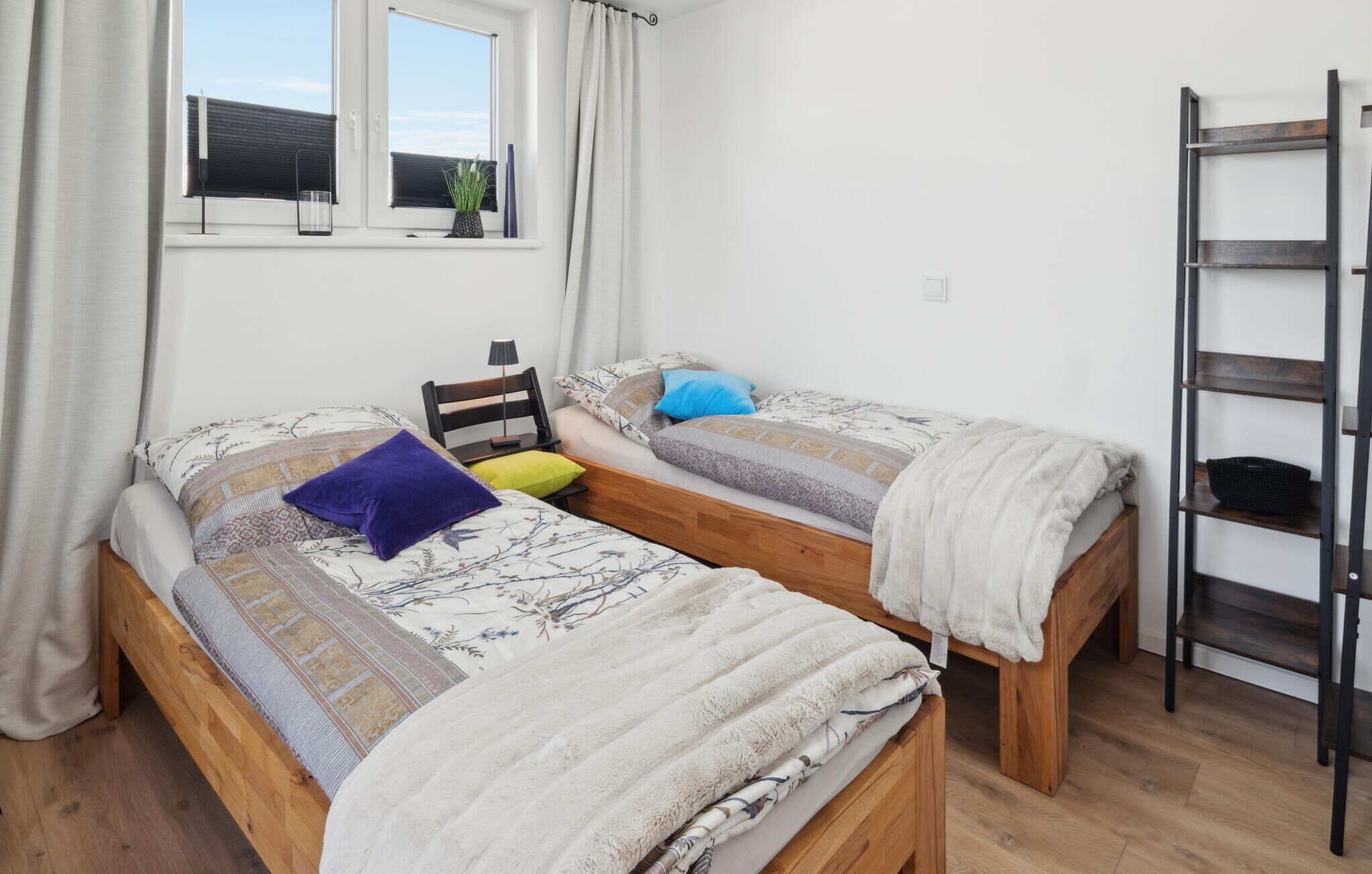 Une chambre avec deux lits en bois, une fenêtre avec des stores, une table de chevet avec une lampe et une échelle en métal appuyée contre le mur.