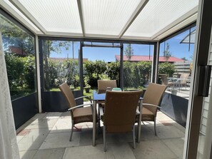 A covered patio with a table and chairs, large windows, and a view of greenery and buildings. - Sint Maartenszee Holiday Home for 4 (Sint Maartensvlotbrug)