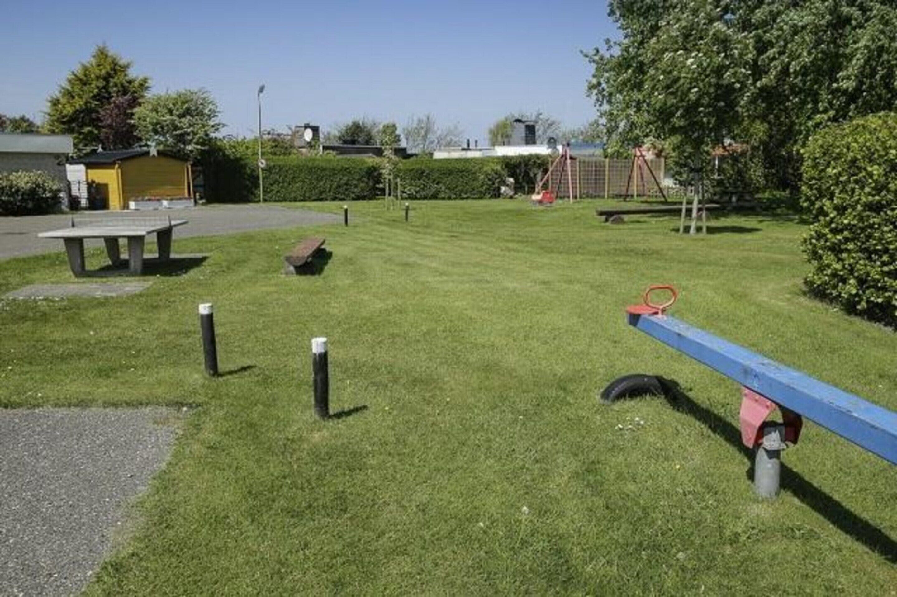 Un área verde bien cuidada con un sube y baja, una pequeña estructura amarilla y un parque infantil con columpios.