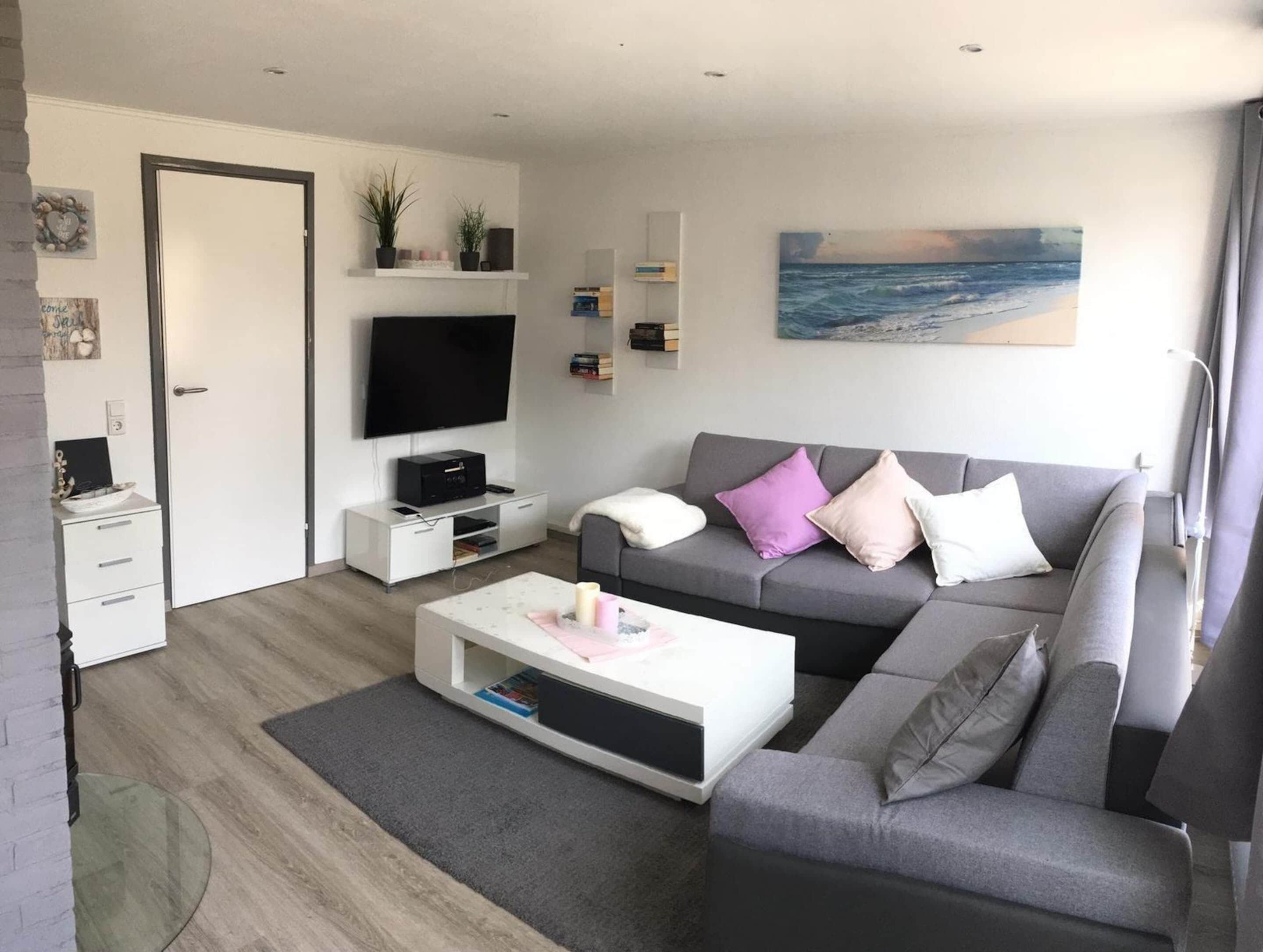 A modern living room with a grey sofa, a flat-screen TV, a white coffee table, and a bookshelf.