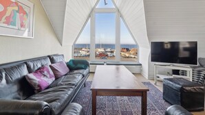 Ein Wohnzimmer mit einer schwarzen Ledersofa, einem Holztisch, einem Flachbildfernseher und einem großen Fenster mit Blick auf Boote und das Meer.