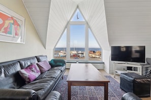 A living room with a black leather sofa, a wooden coffee table, a flat-screen TV, and a large window offering a view of boats and the sea.