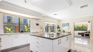 Een moderne keuken met witte kasten, een granieten aanrecht en een groot schiereiland. Er zijn ramen met uitzicht op groen en een zithoek met een bank en tafel.