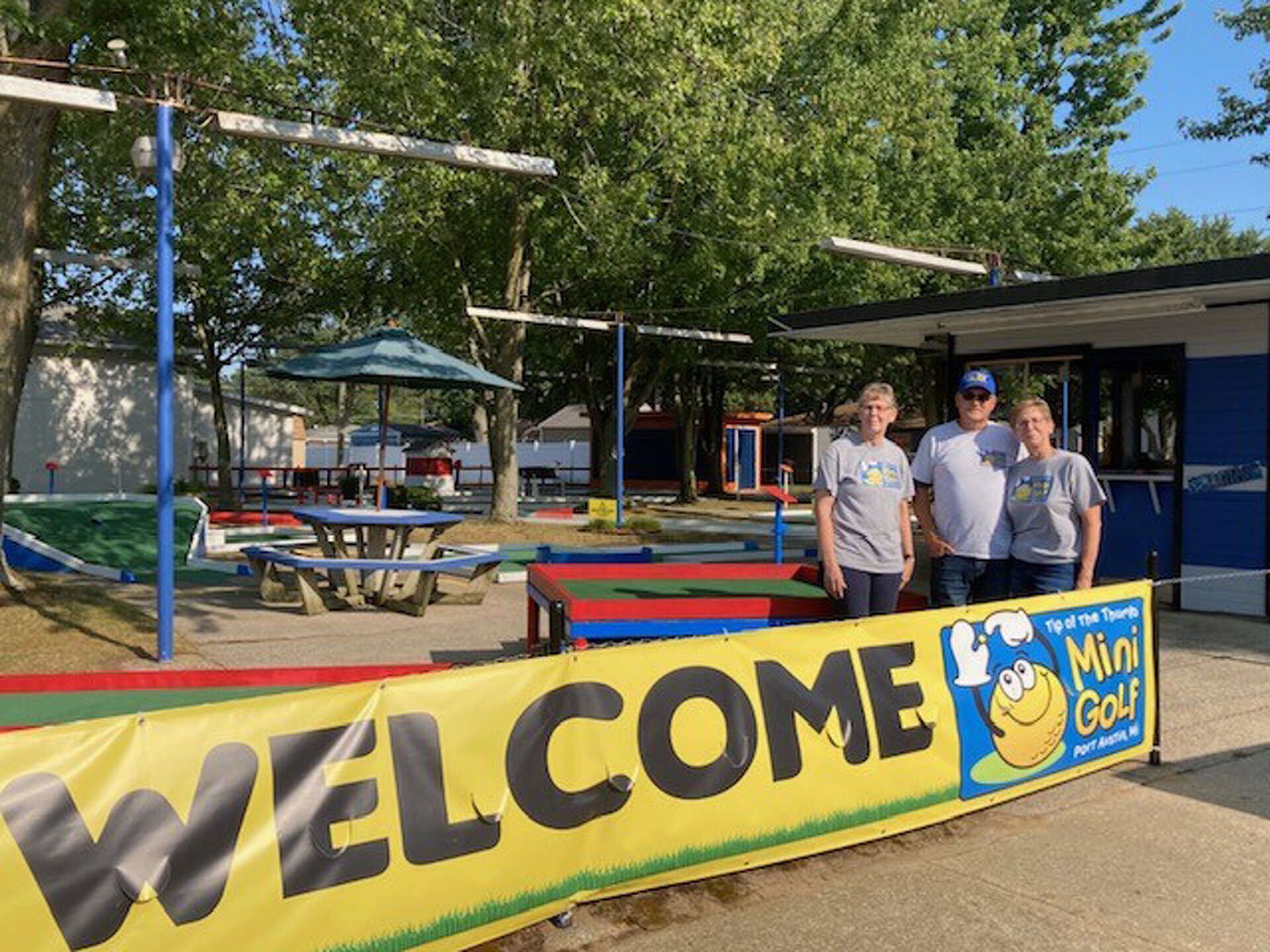 En banner med teksten "WELCOME" og "Mini Golf", et picnicbord og et legeareal.