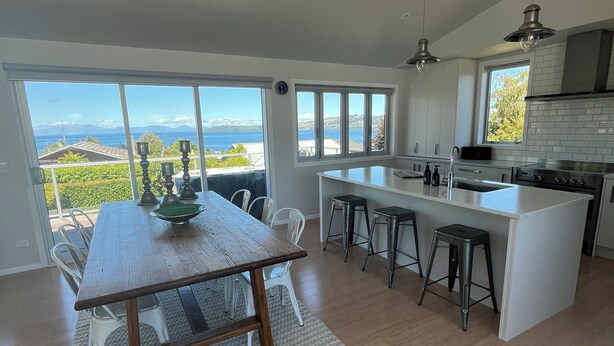A modern kitchen with a large island, bar stools, and a dining area with a view of the ocean. - HILLTOP, CENTRAL, QUIET, LOCATION WITH STUNNING LAKE AND MOUNTAIN VIEWS. (Taupō)