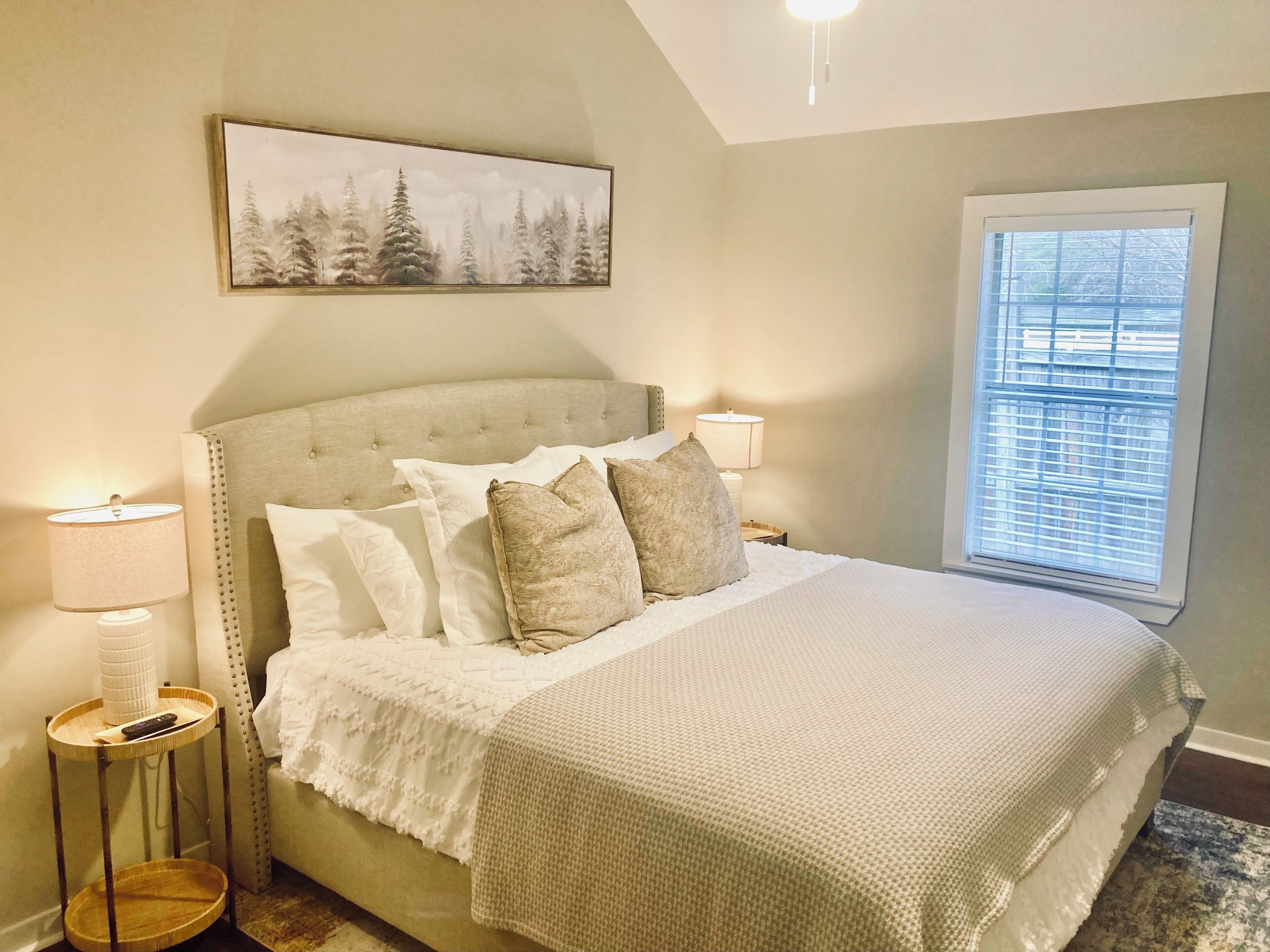 Une chambre à coucher avec un grand lit, une tête de lit, des lampes de chevet, une fenêtre avec des stores et un tableau au mur.