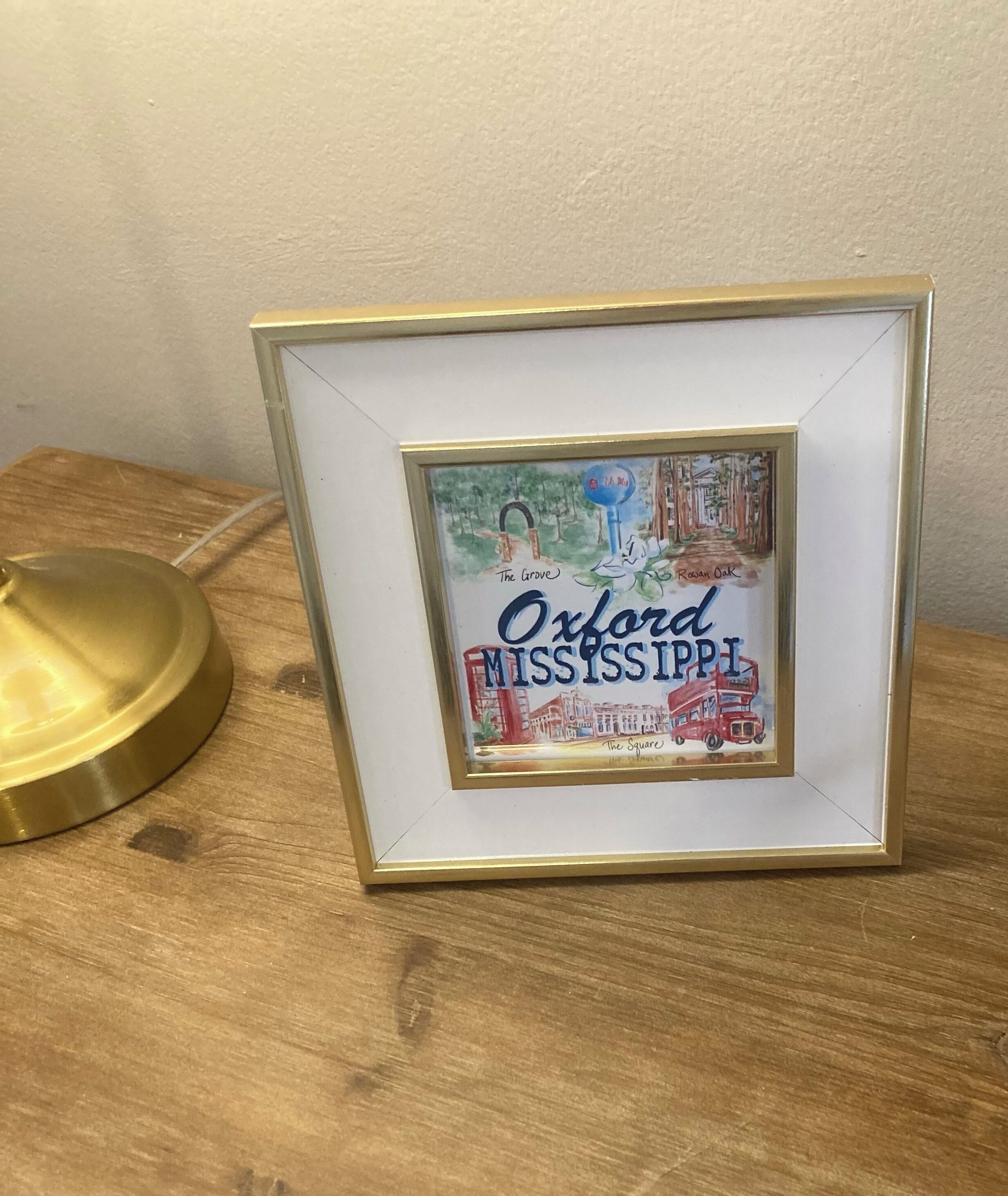 A framed picture of Oxford, Mississippi, with a gold frame and a brass lamp beside it.