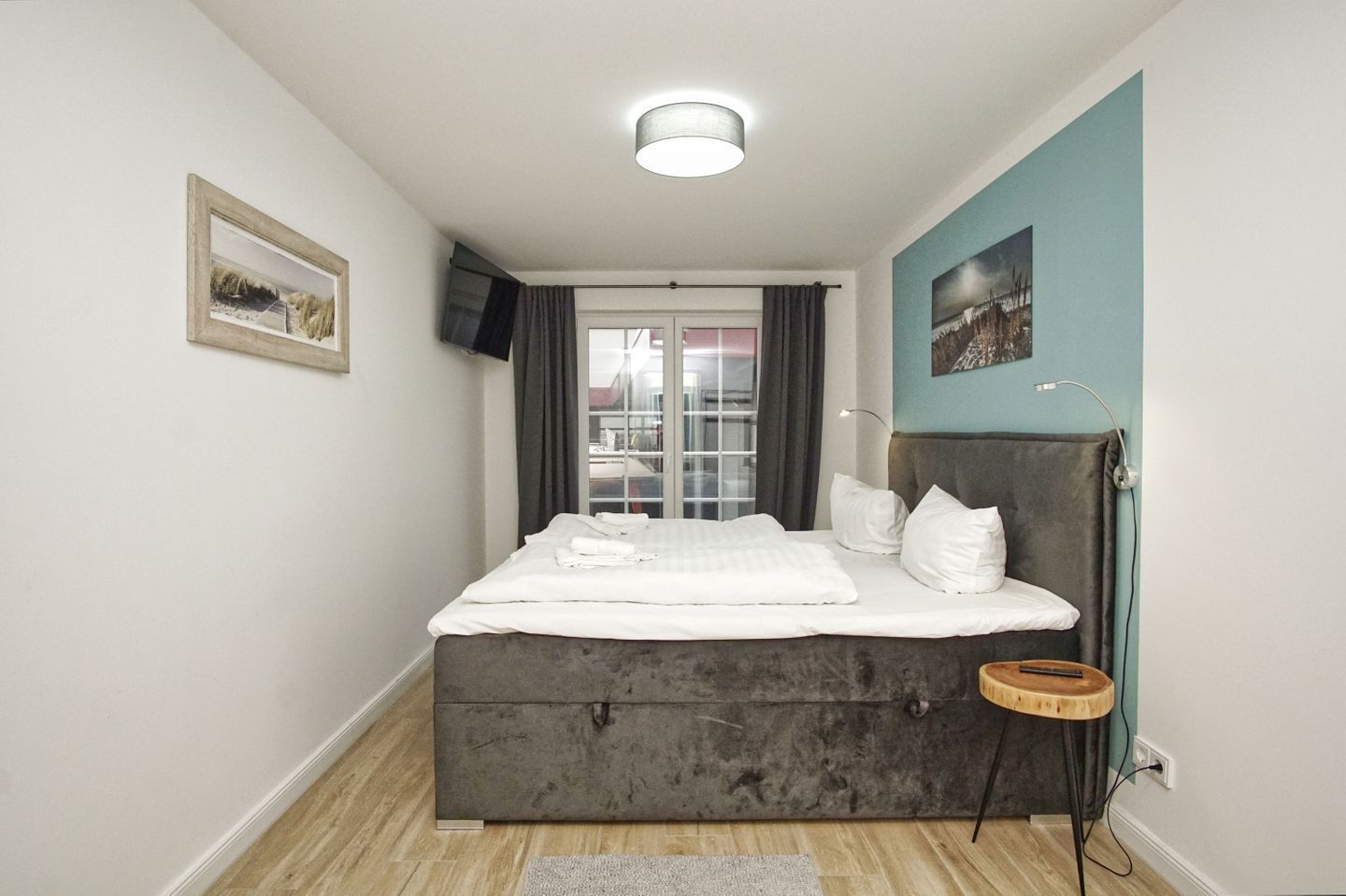 Ein modernes Schlafzimmer mit einem großen Bett, einem kleinen Nachttisch, einem an der Wand montierten Fernseher und einem Fenster mit Vorhängen.