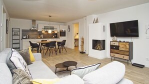 Uma sala de estar moderna com sofá, uma área de jantar com cadeiras, cozinha com armários, lareira e TV na parede.