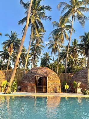Un espace aménagé au bord de la piscine, avec des cabanes à toit de chaume, des palmiers et un ciel d’un bleu limpide.