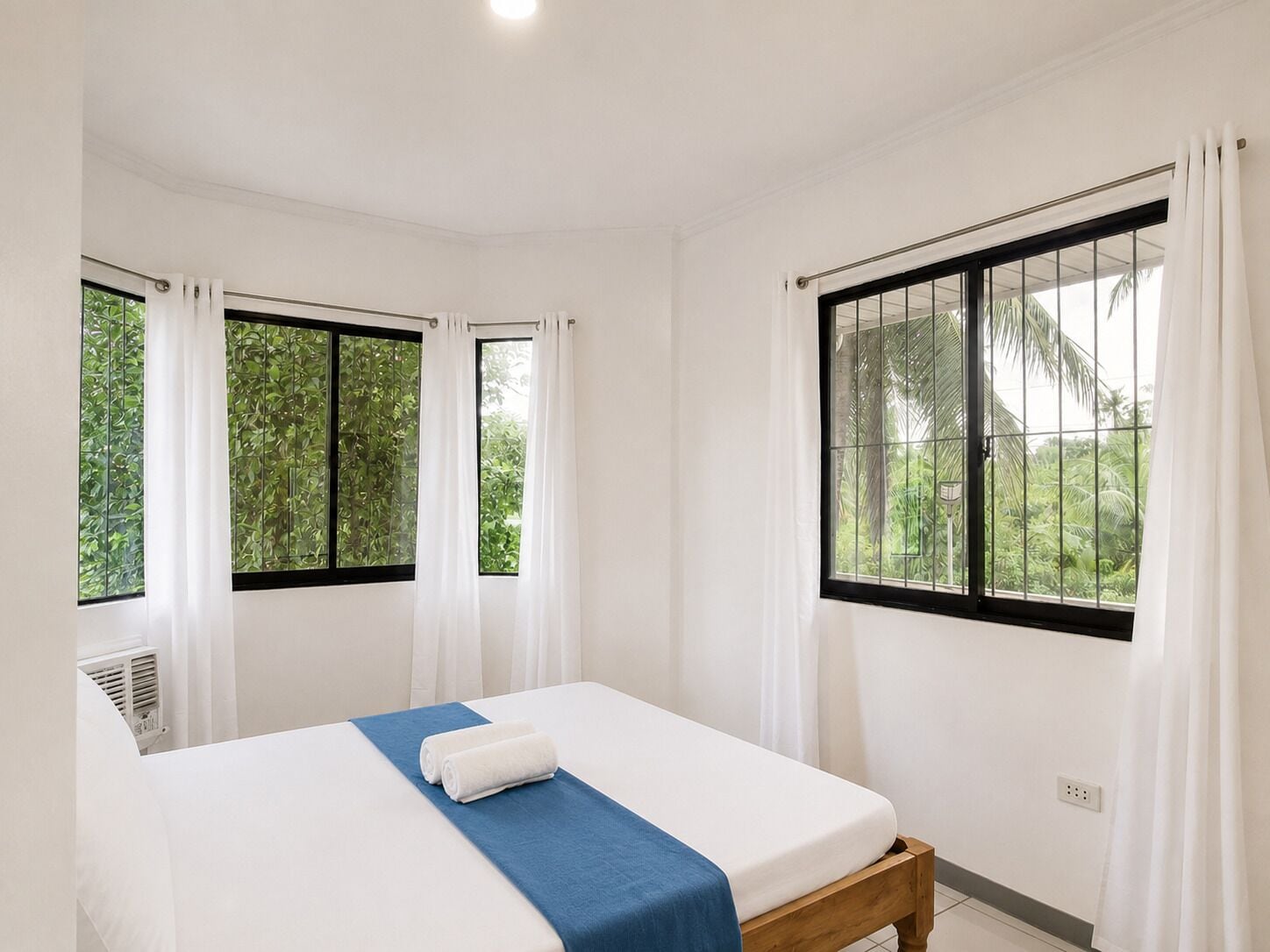 A room with a bed, white curtains, and windows with a view of greenery.