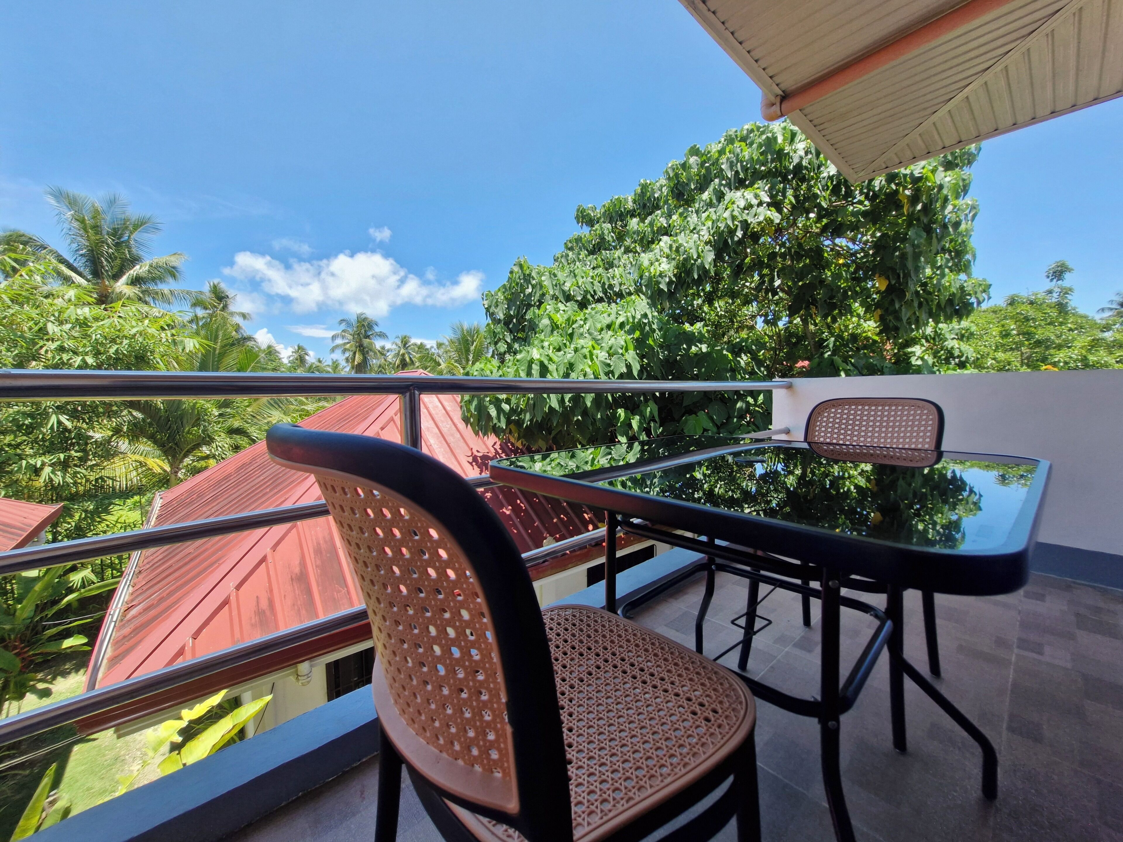 Un balcon avec une table et des chaises, une paroi en verre et une vue sur la verdure et les bâtiments.
