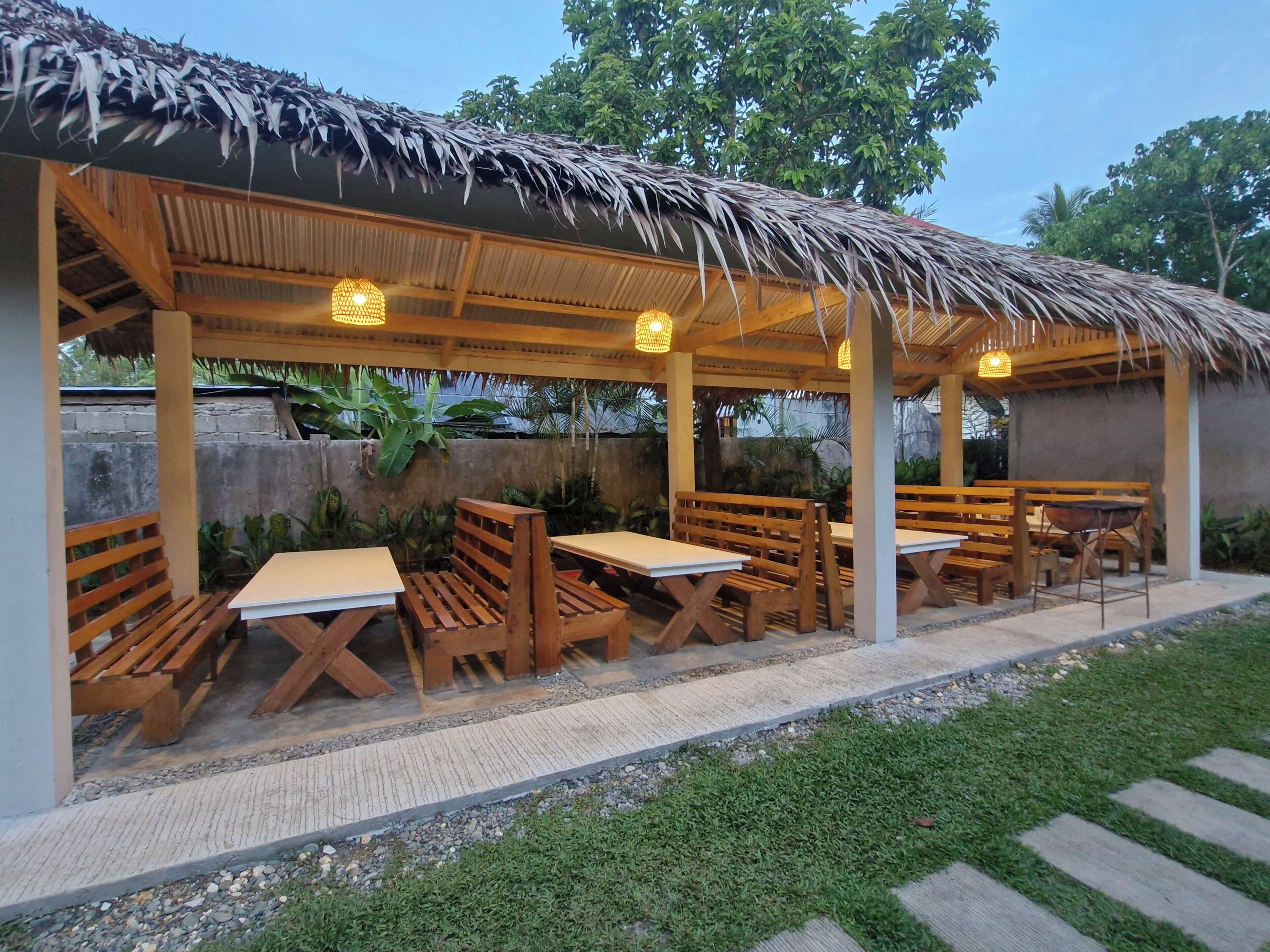 An outdoor patio with wooden benches and tables, a thatched roof, and hanging lights.