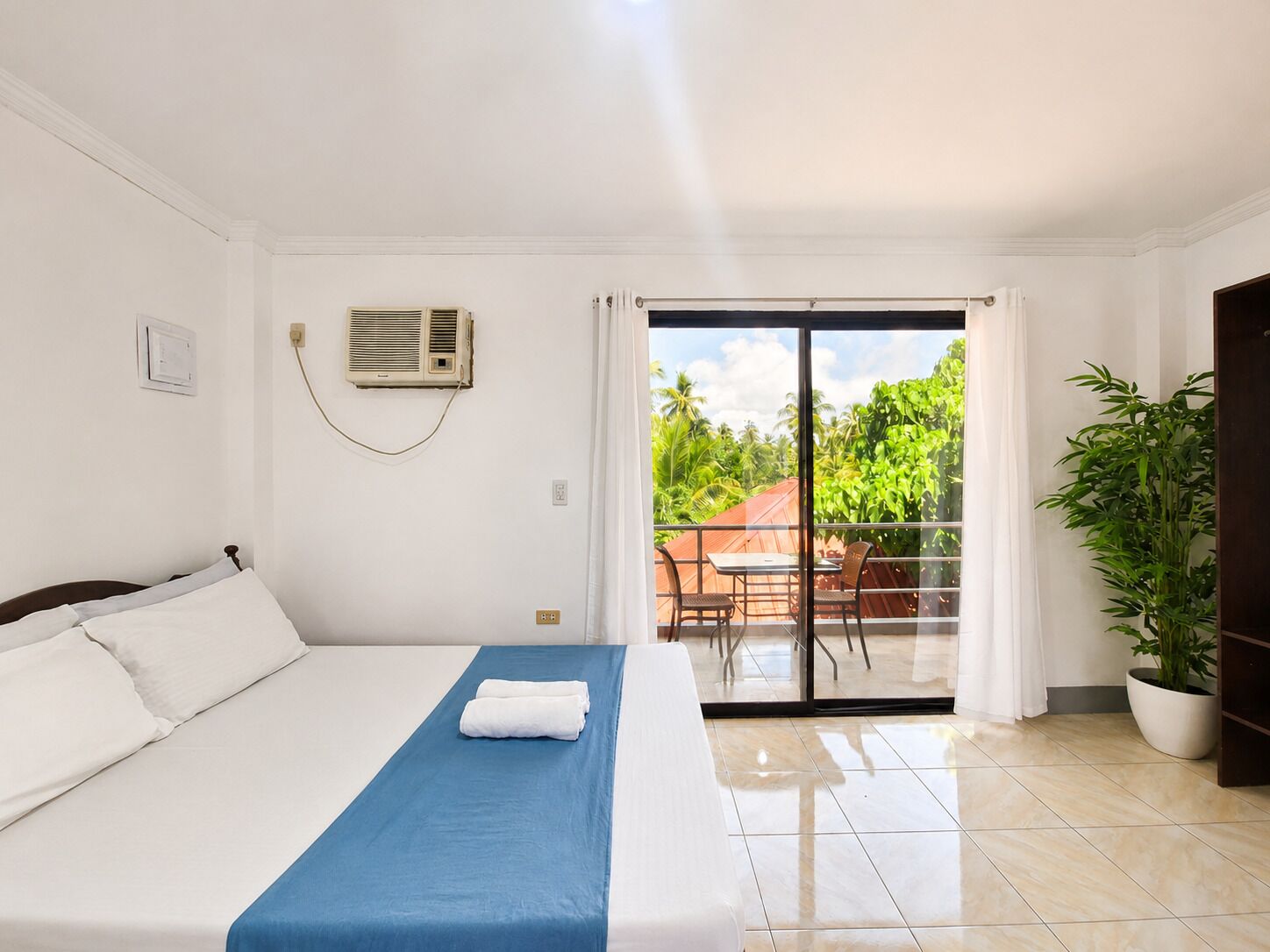 Una habitación de hotel con cama, una colcha azul, un equipo de aire acondicionado, una puerta corrediza de vidrio a un balcón y una planta en maceta.