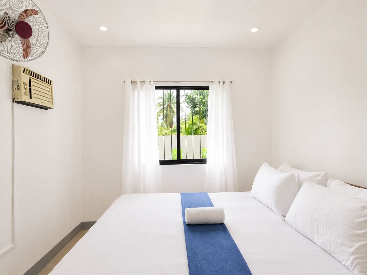Una habitación de hotel con una cama, un ventanal con cortinas, un ventilador de pared y un termostato.