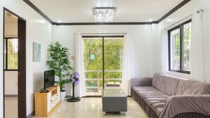 Una sala de estar moderna con un sofá, una mesa de centro, un televisor sobre un soporte de madera, una planta en maceta y un amplio ventanal con vistas a la vegetación.