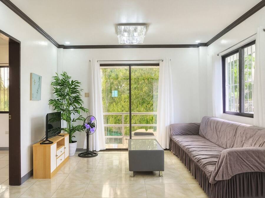 A modern living room with a sofa, a coffee table, a TV on a wooden stand, a potted plant, and a large window with a view of greenery.