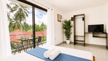 A hotel room with a bed, a TV, a potted plant, a balcony with a table and chairs, and a view of palm trees.