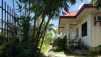 Un sendero conduce a una casa con techo rojo, rodeada de abundante vegetación y una cerca de bambú.