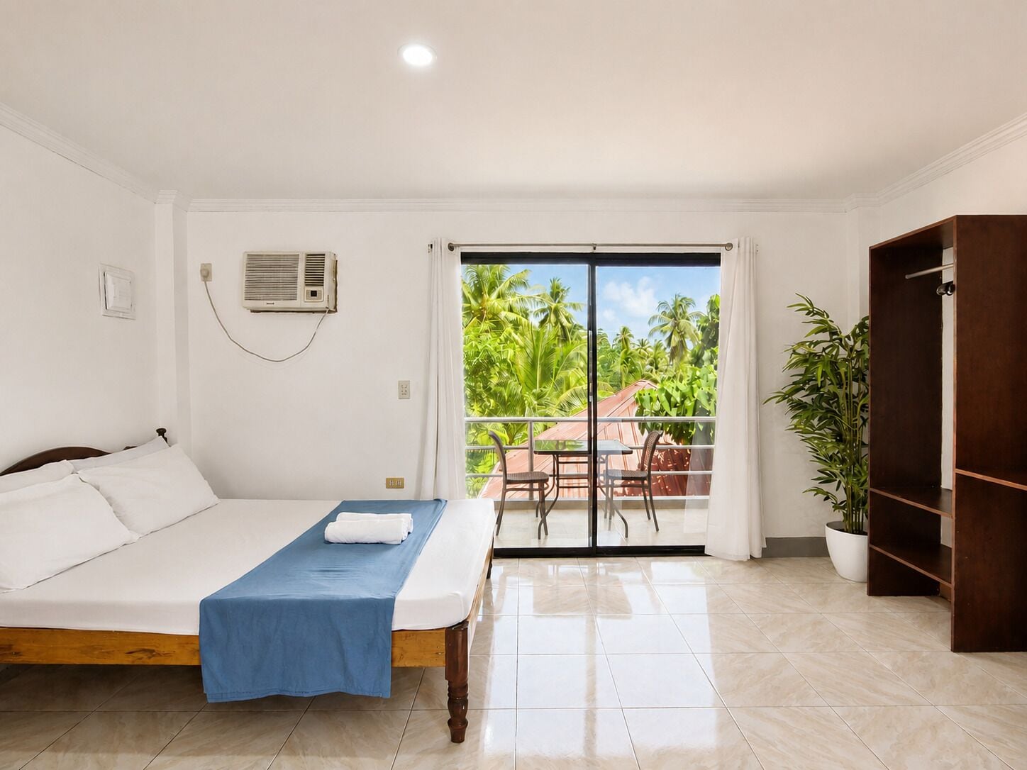 Una camera d'albergo con un letto, una coperta blu, una porta scorrevole in vetro che dà su un balcone, una pianta in vaso e un armadio in legno.