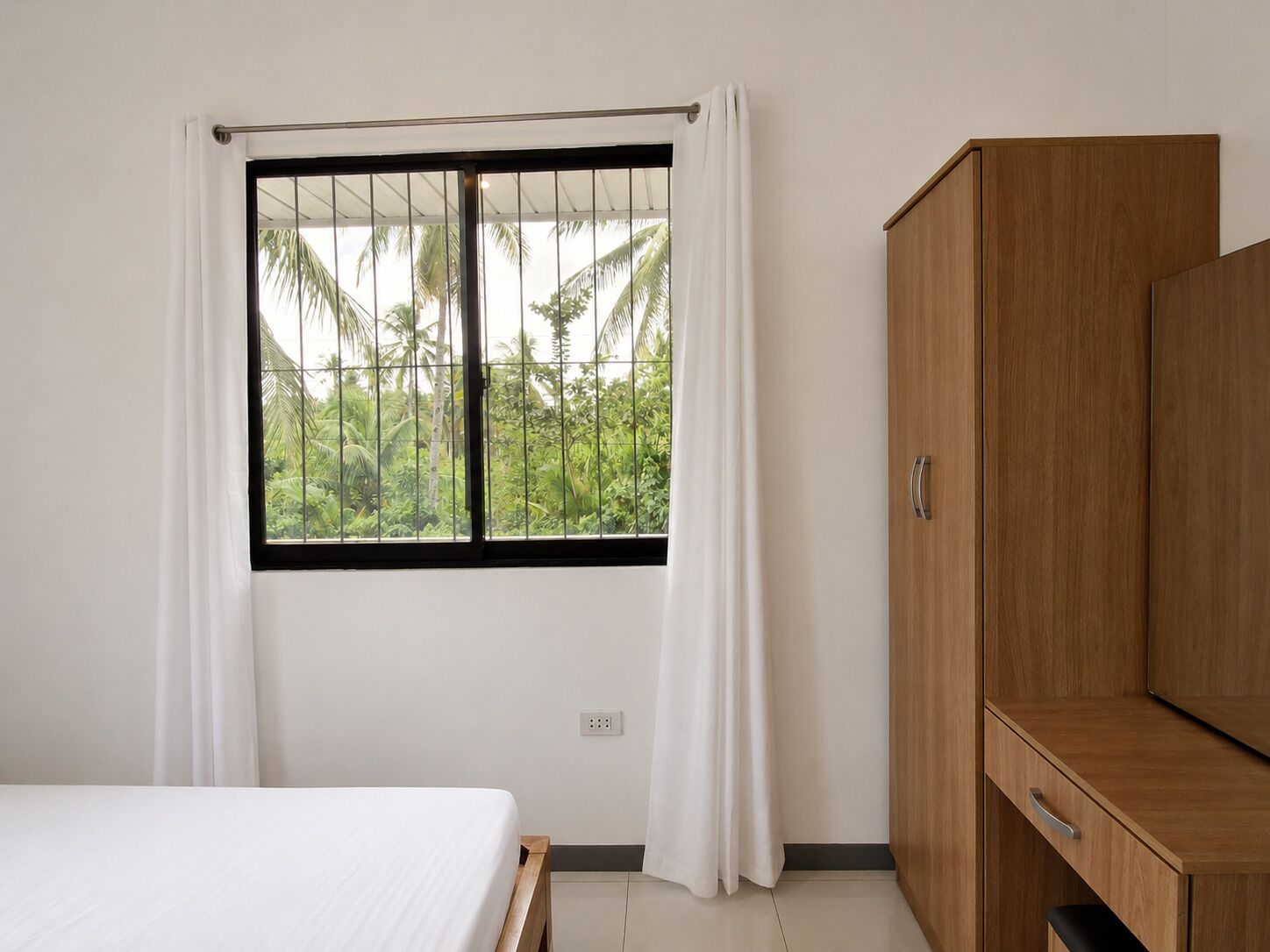A room with a bed, a window with curtains, a wardrobe, and a view of greenery outside.