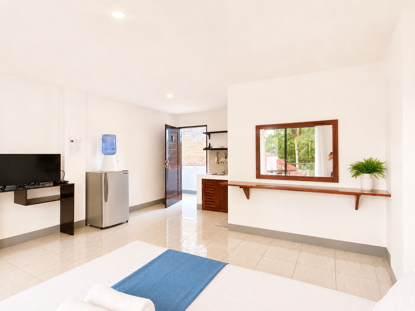 Habitación de hotel con televisión de pantalla plana, dispensador de agua, refrigerador, estante de madera con planta y ventana con vista a vegetación.