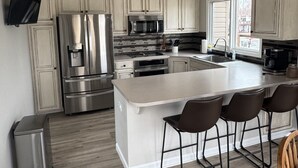 A modern kitchen with white cabinets, stainless steel appliances, a central island with bar stools, and a flat-screen TV mounted on the wall.