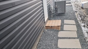 Ein Weg, der zu einem Gebäude mit grauer Fassade und einer kleinen Holterrasse führt.