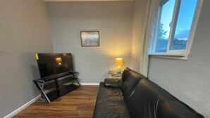 Un salon avec un canapé en cuir noir, un téléviseur posé sur un meuble, une lampe et une fenêtre.