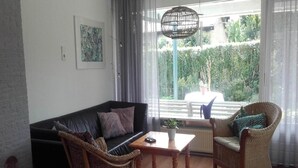 Un salon avec un canapé en cuir noir, deux fauteuils en osier, une table basse en bois et une vue sur un balcon doté d’une balustrade et de plantes à l’extérieur.