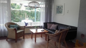 Un salon avec un canapé en cuir noir, des fauteuils en osier, une table basse en bois et une vue sur la verdure grâce à de grandes fenêtres.