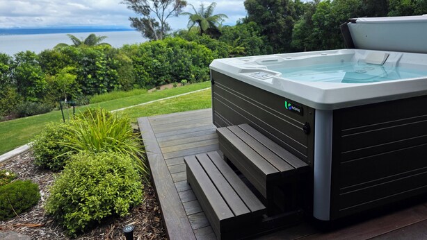 Un jacuzzi con panel de control, rodeado por una terraza de madera y abundante vegetación, con vistas a un cuerpo de agua.