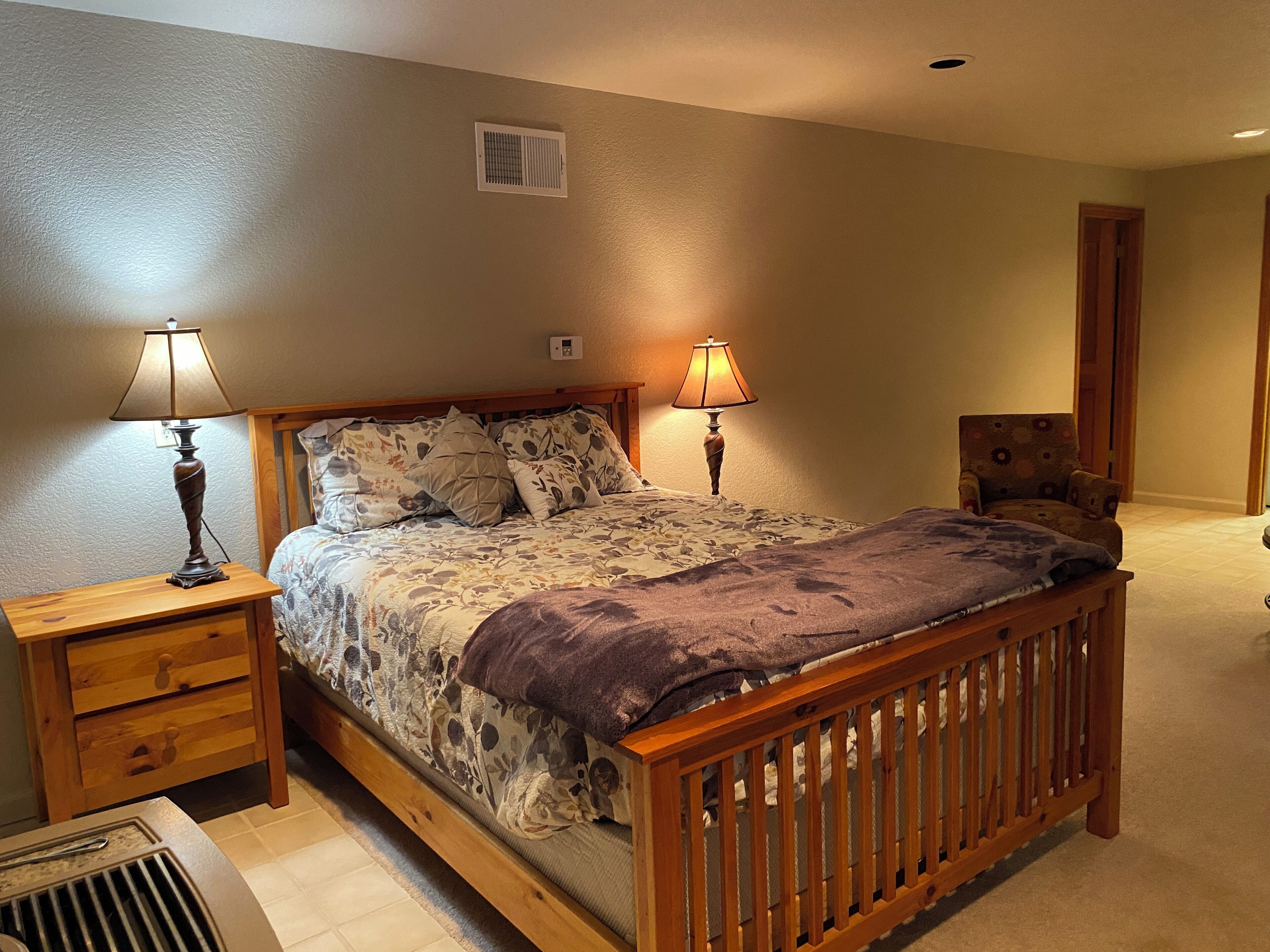 A bedroom with a wooden bed, two bedside tables with lamps, a chair, and a vent on the wall.
