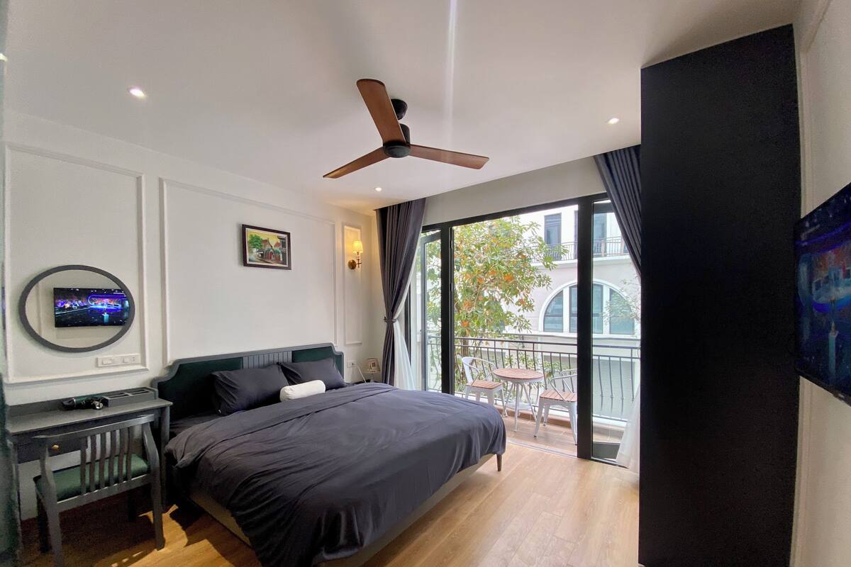 A modern hotel room with a large bed, a ceiling fan, a TV mounted on the wall, and a balcony with a view of the city.