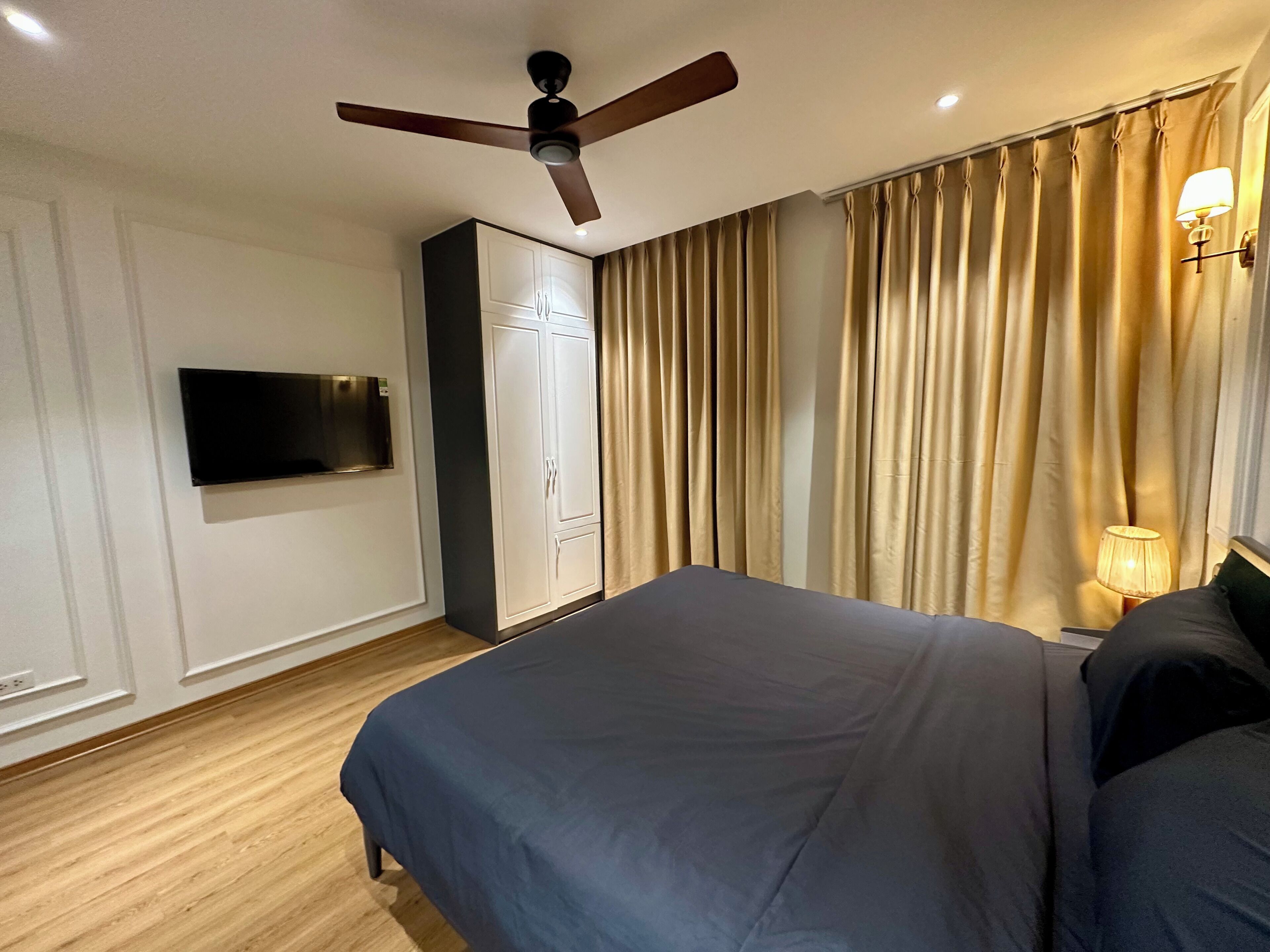 A hotel room with a bed, a ceiling fan, a TV mounted on the wall, and a wardrobe.