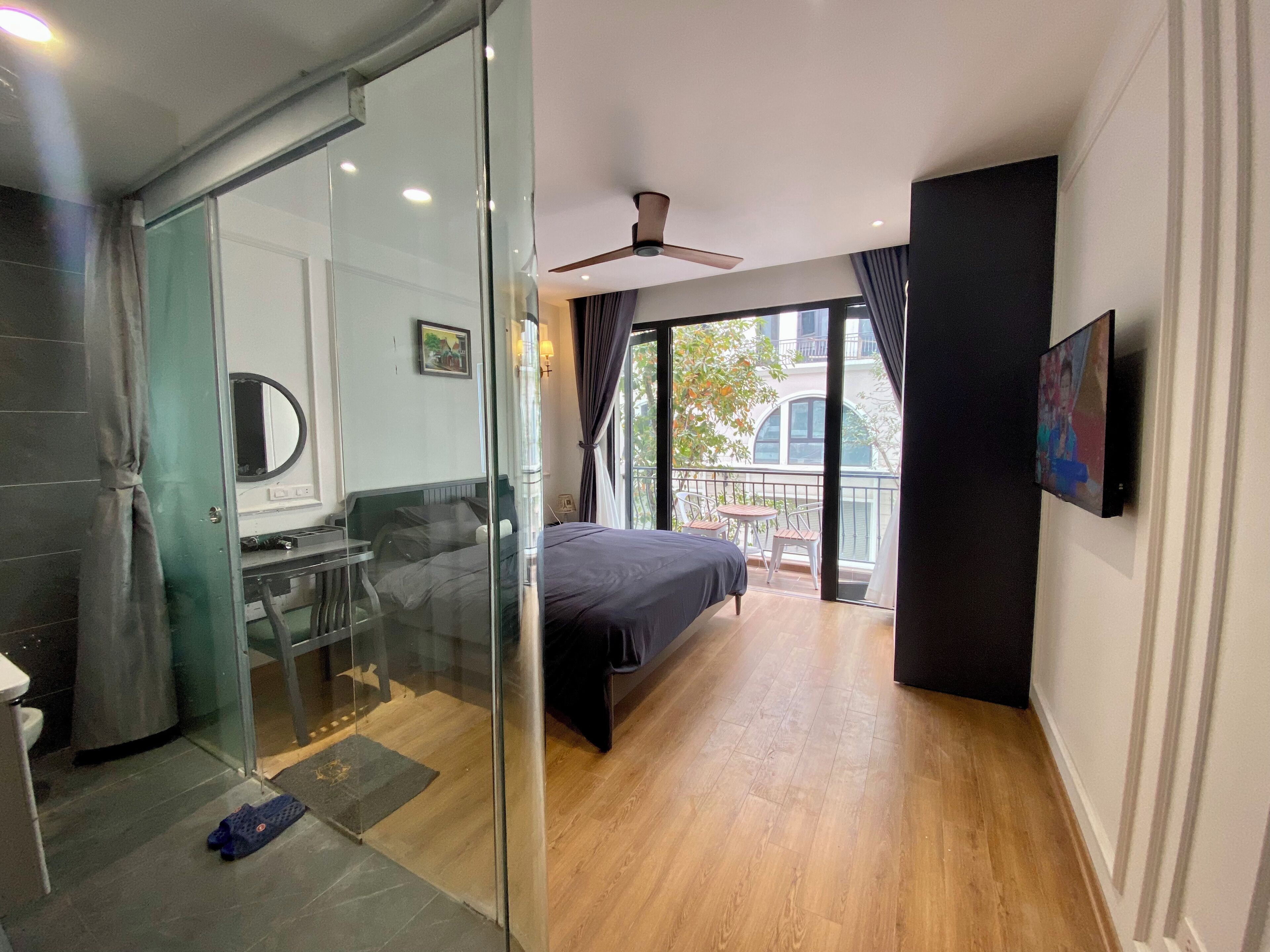 A modern hotel room with a glass partition, a bed, a small balcony with a table and chairs, and a ceiling fan.